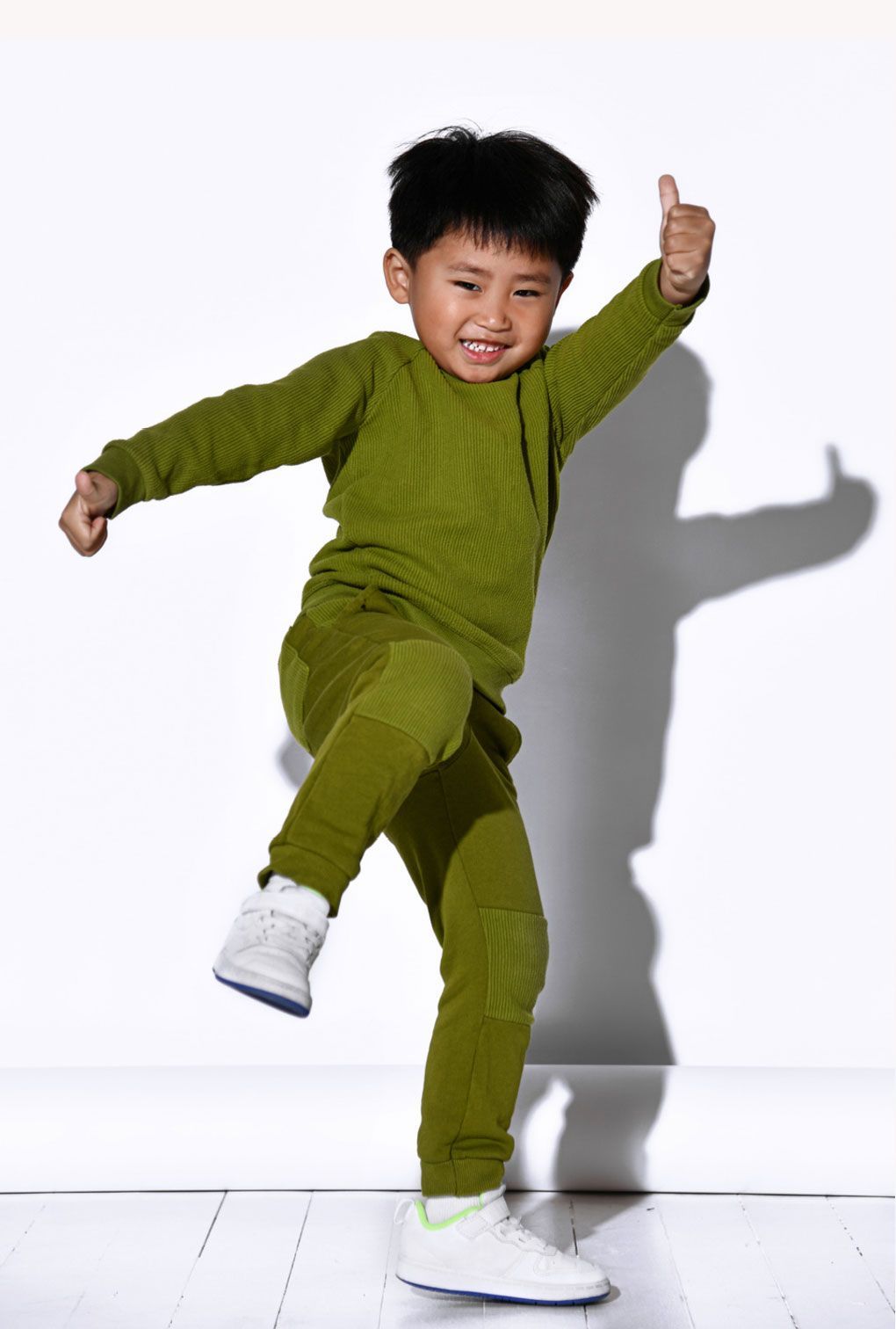 A young boy in a green sweater and pants is giving a thumbs up.