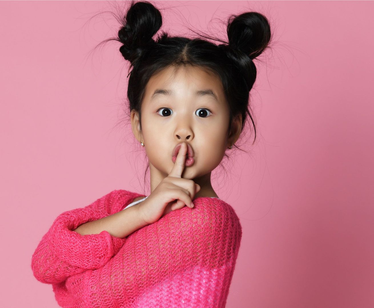 A little girl in a pink sweater is holding her finger to her mouth.