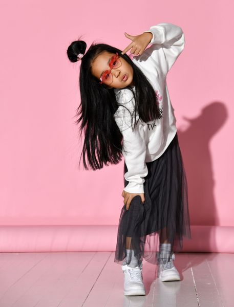 Young girl in sunglasses and white sweatshirt, making a gun hand gesture, leaning to the side. Pink background.