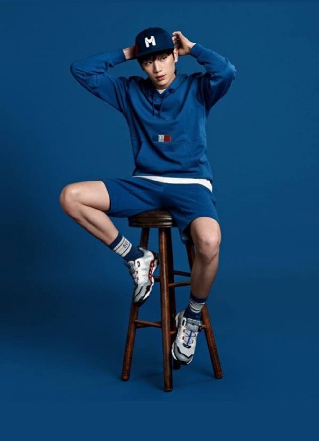 Man in blue outfit sits on a stool, posing, with a blue background.