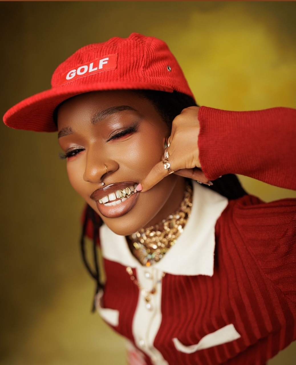 Woman in red cap and top winks, makes phone gesture, gold jewelry, and bright smile.