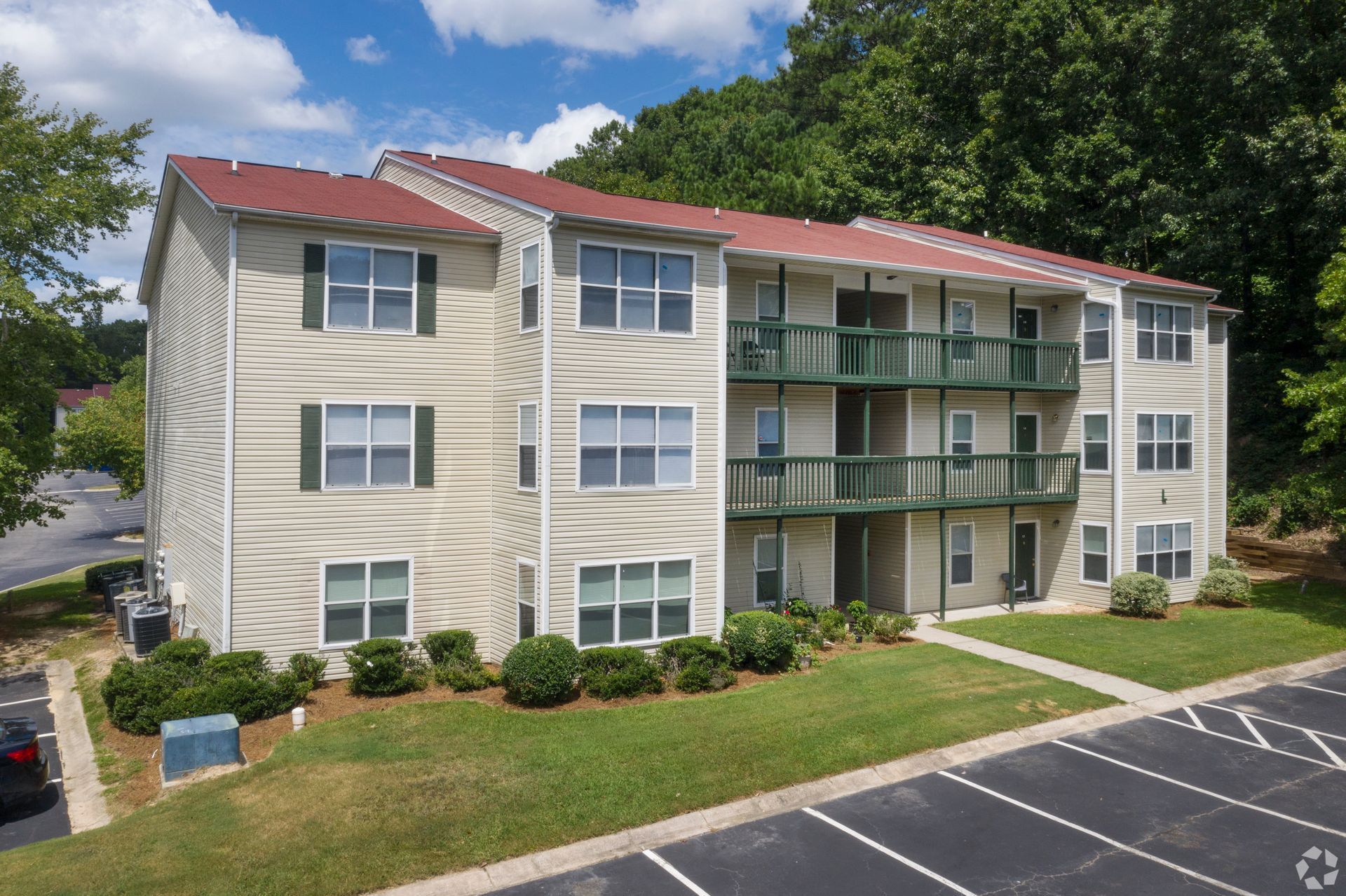 A three-story, beige apartment building with a red roof, green balconies, and an adjacent parking lot.