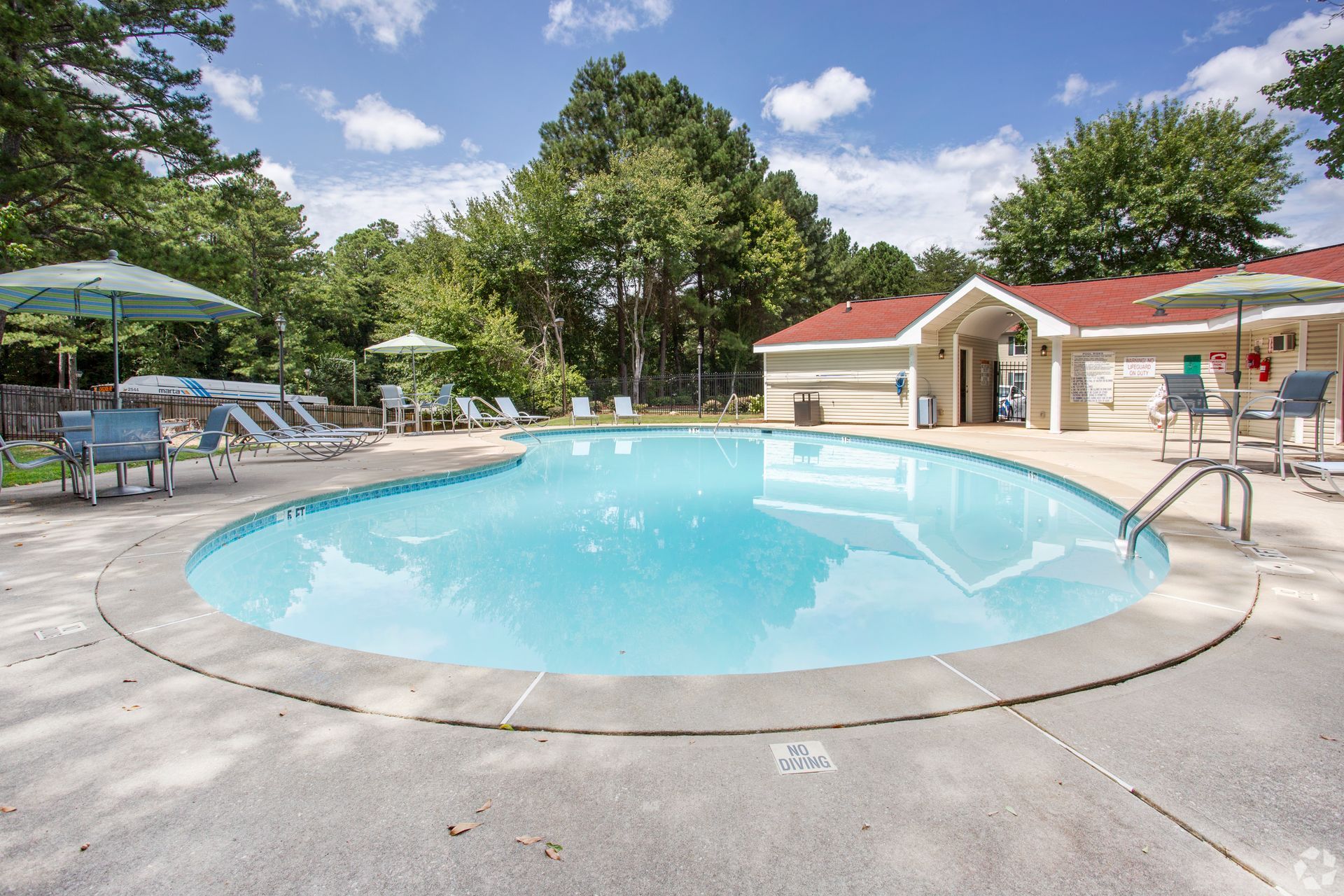 An outdoor swimming pool surrounded by a concrete deck, lounge chairs with umbrellas, and a small building with a red roof.