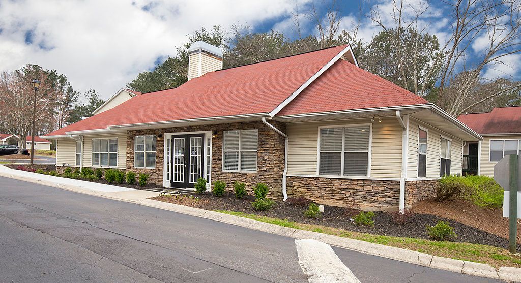 Single-story community center with a red roof, stone facade, and dark double doors, situated beside a paved road.