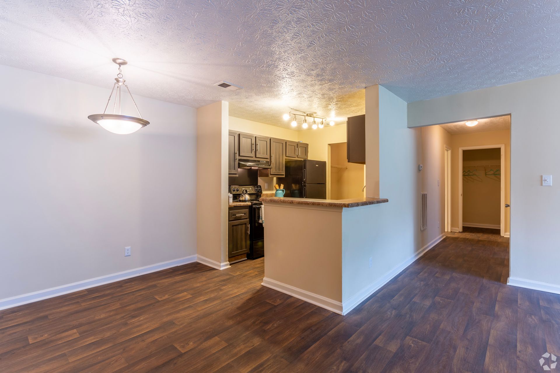 A kitchen with dark cabinets and a breakfast bar opens into a dining area with wood-look flooring and a pendant light.