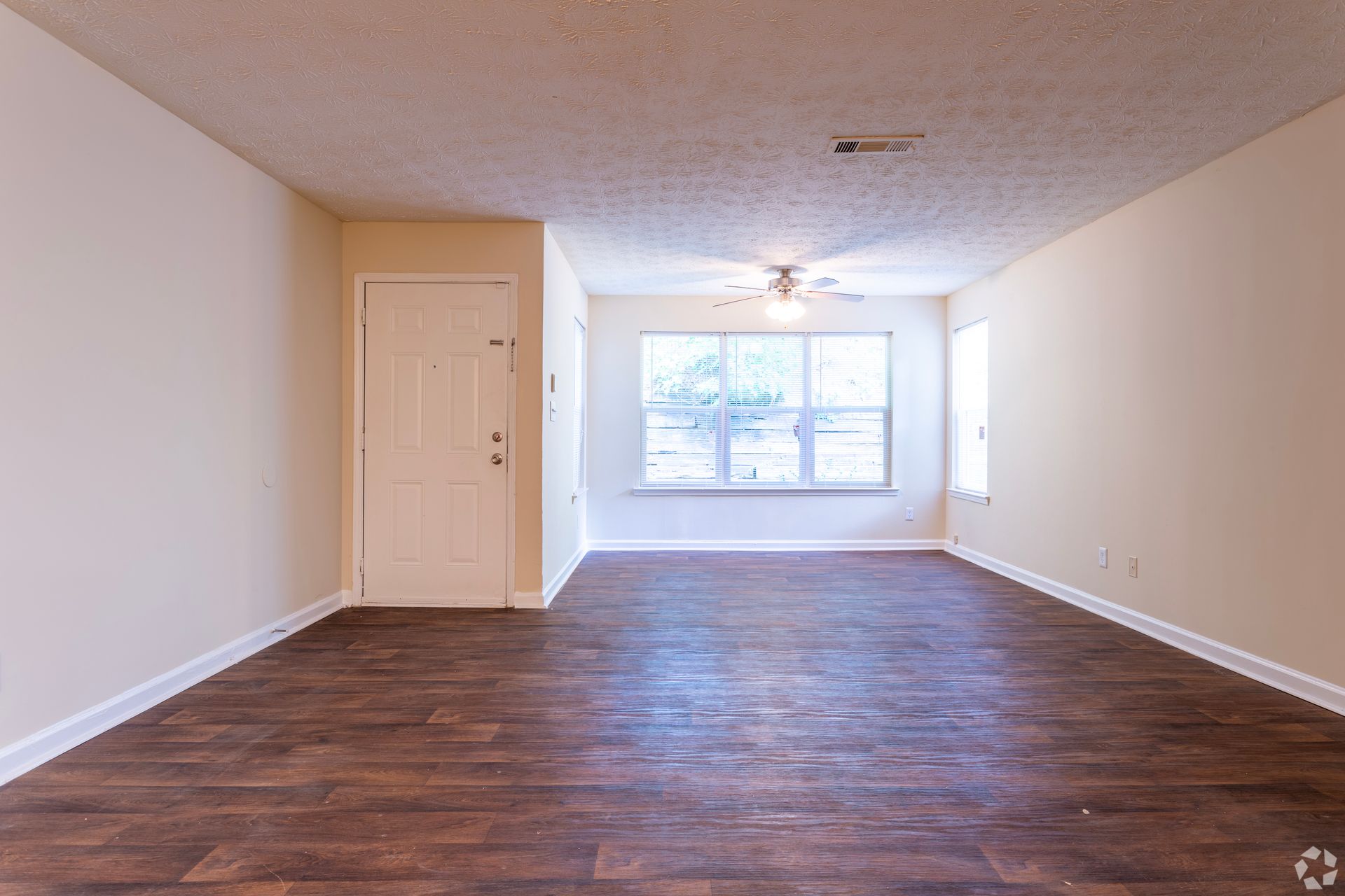 An empty room with light tan walls, dark wood-look floors, a white front door, and a window with a ceiling fan overhead.