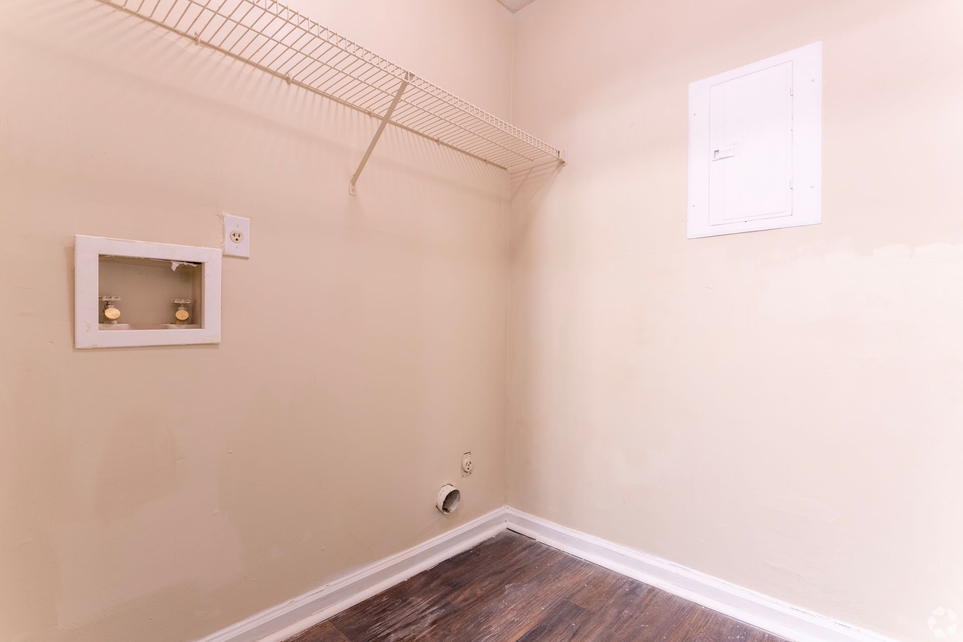 A laundry room corner with beige walls, wood-look flooring, a wire shelf, plumbing hookups, and a white access panel.