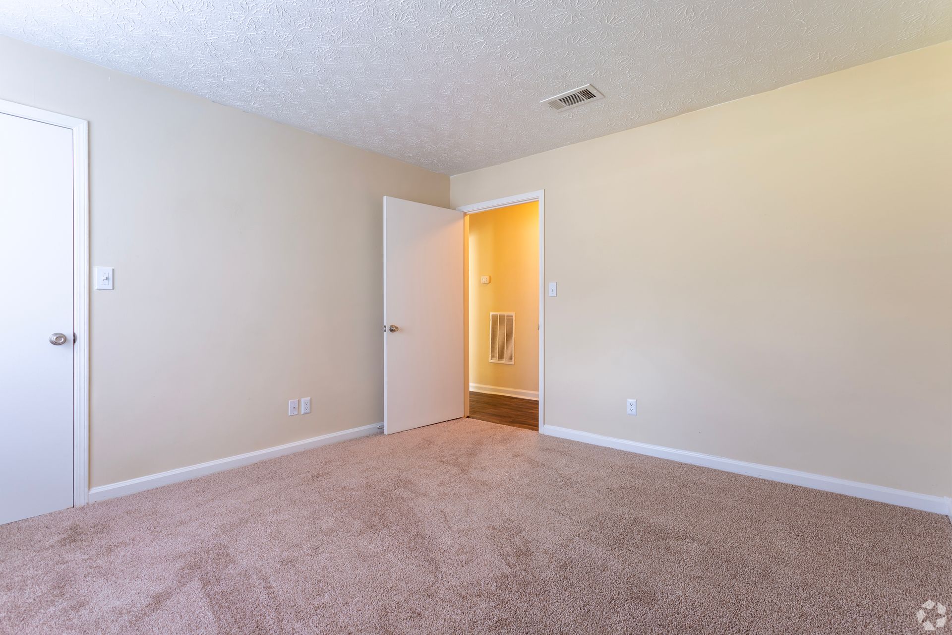 An empty room with tan walls and beige carpet, featuring a closet door, an open doorway, and a popcorn ceiling.