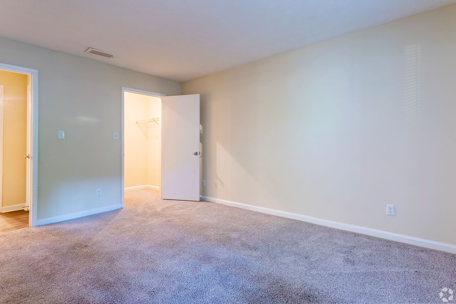 An empty, light-walled bedroom with beige carpeting, featuring an open doorway leading to a walk-in closet.