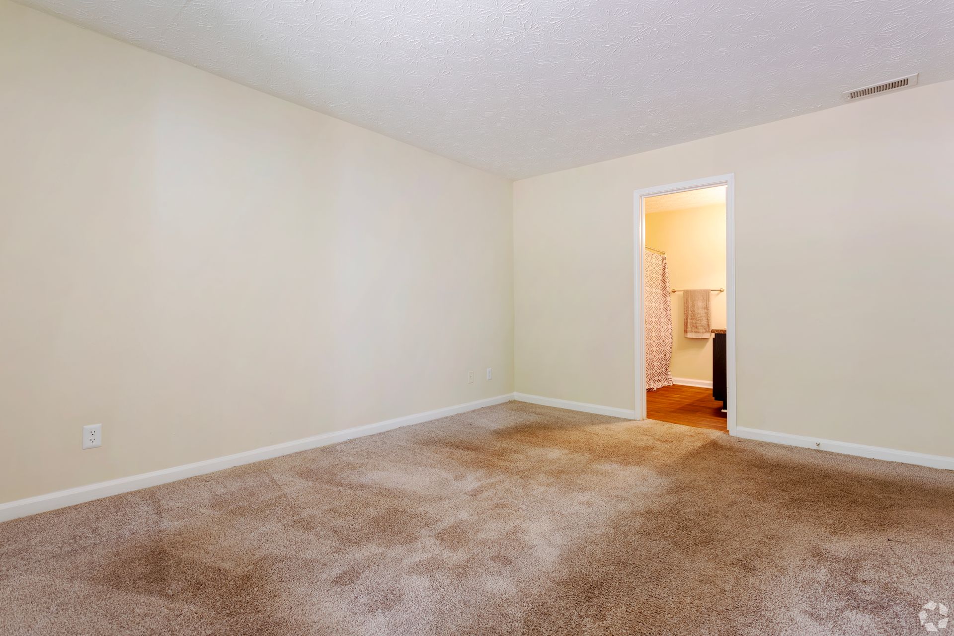 An empty room with light beige walls and tan carpet, featuring a doorway leading to a bathroom.