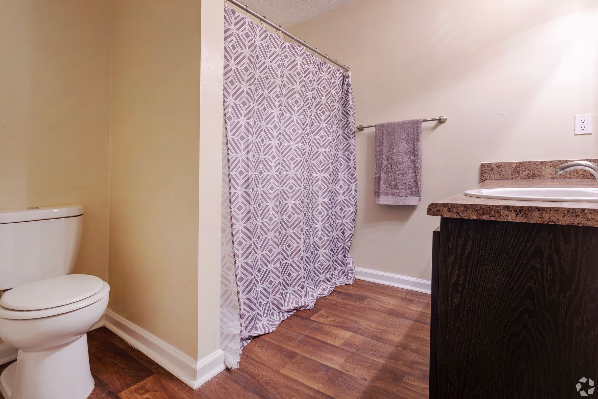 A bathroom with a white toilet, dark wood vanity, a patterned shower curtain, and a grey towel hanging on the wall.