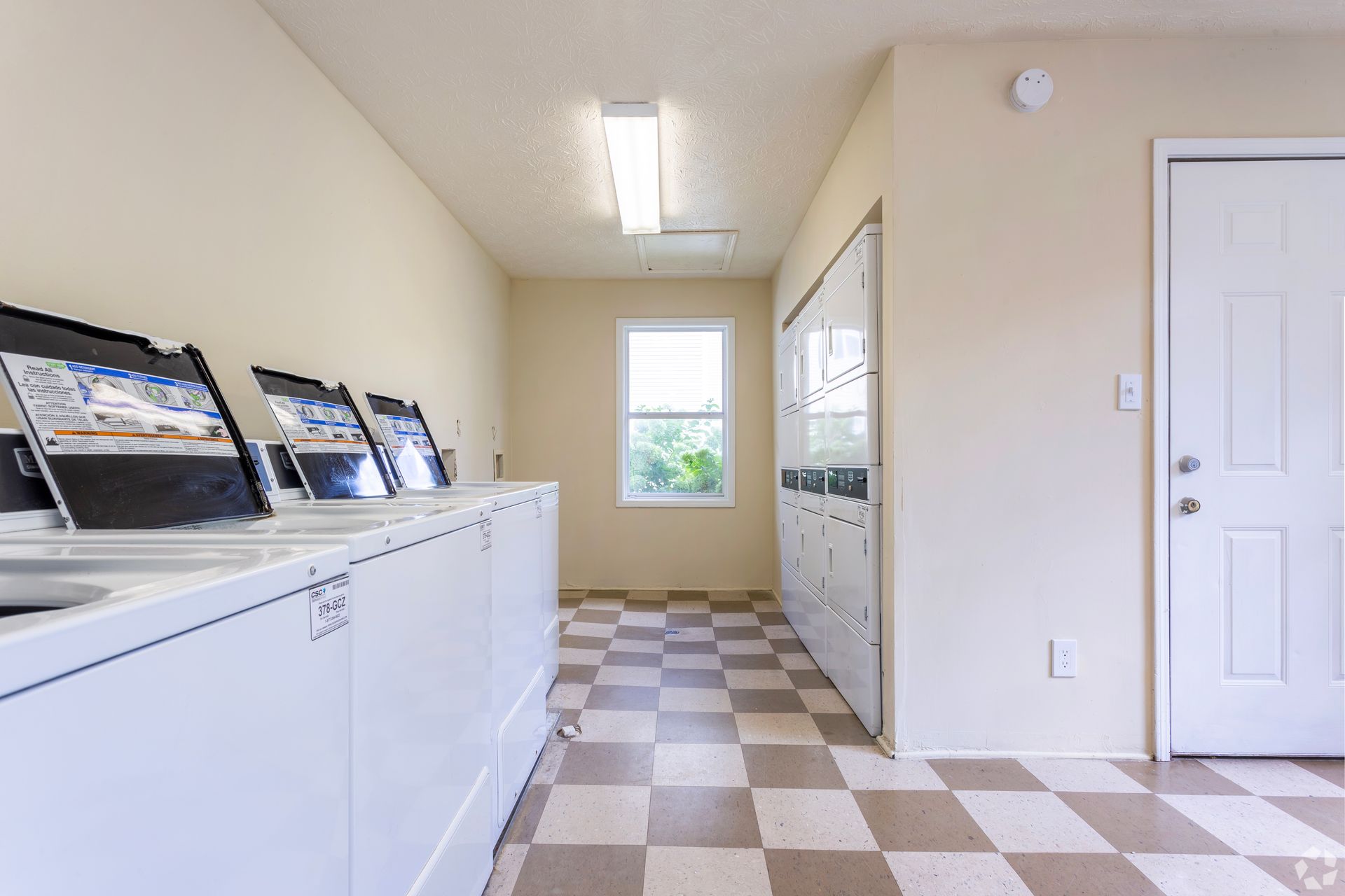 A bright laundry room with white machines, checkered tile flooring, a window, and a door on the right.
