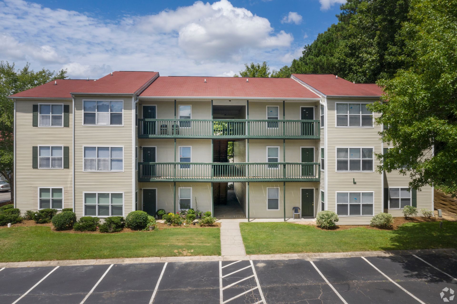Three-story apartment building with tan siding, a red gabled roof, green balconies, and an asphalt parking lot in front.