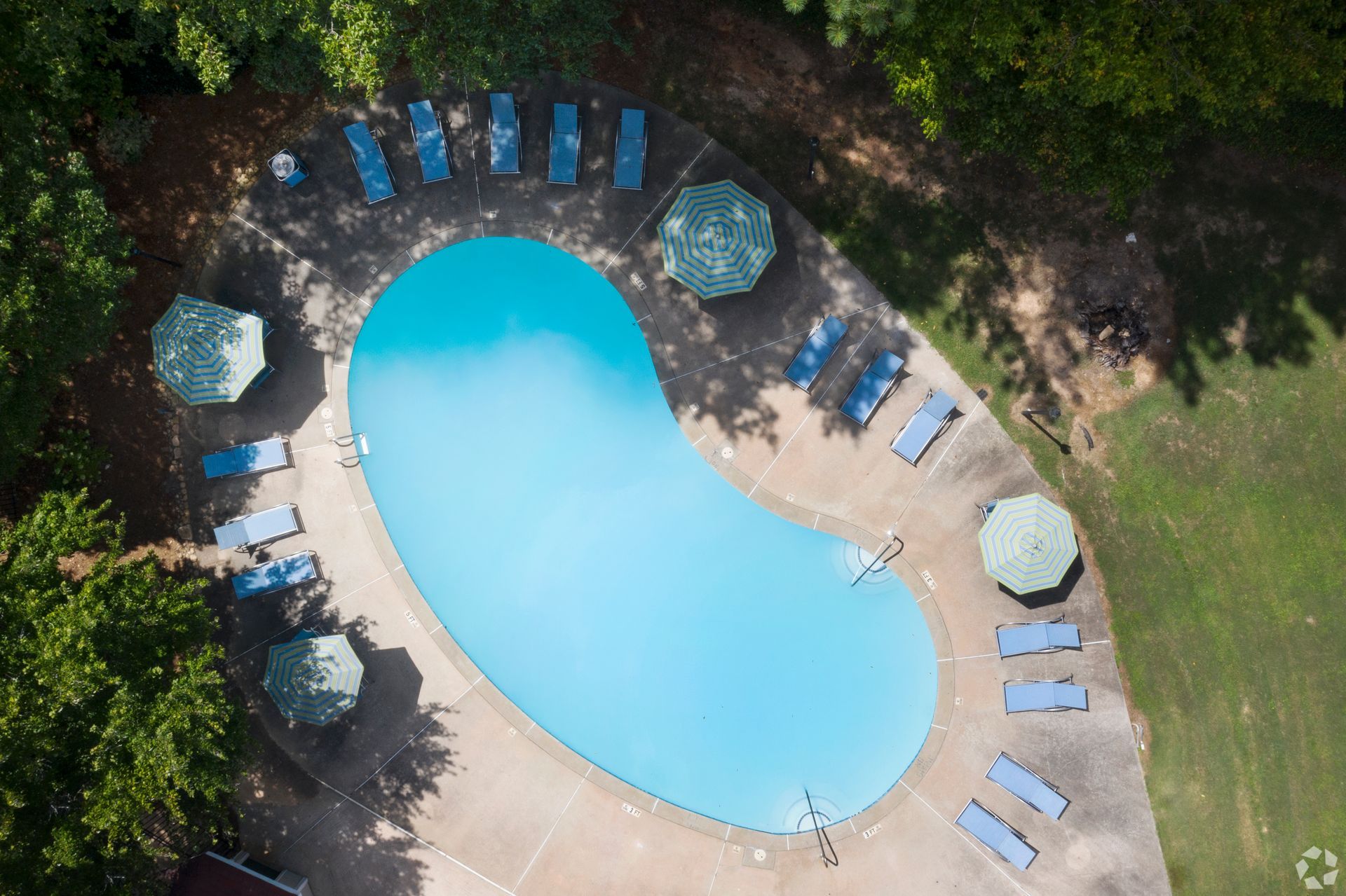 Aerial view of a kidney-shaped swimming pool surrounded by lounge chairs and umbrellas on a paved deck in a wooded area.