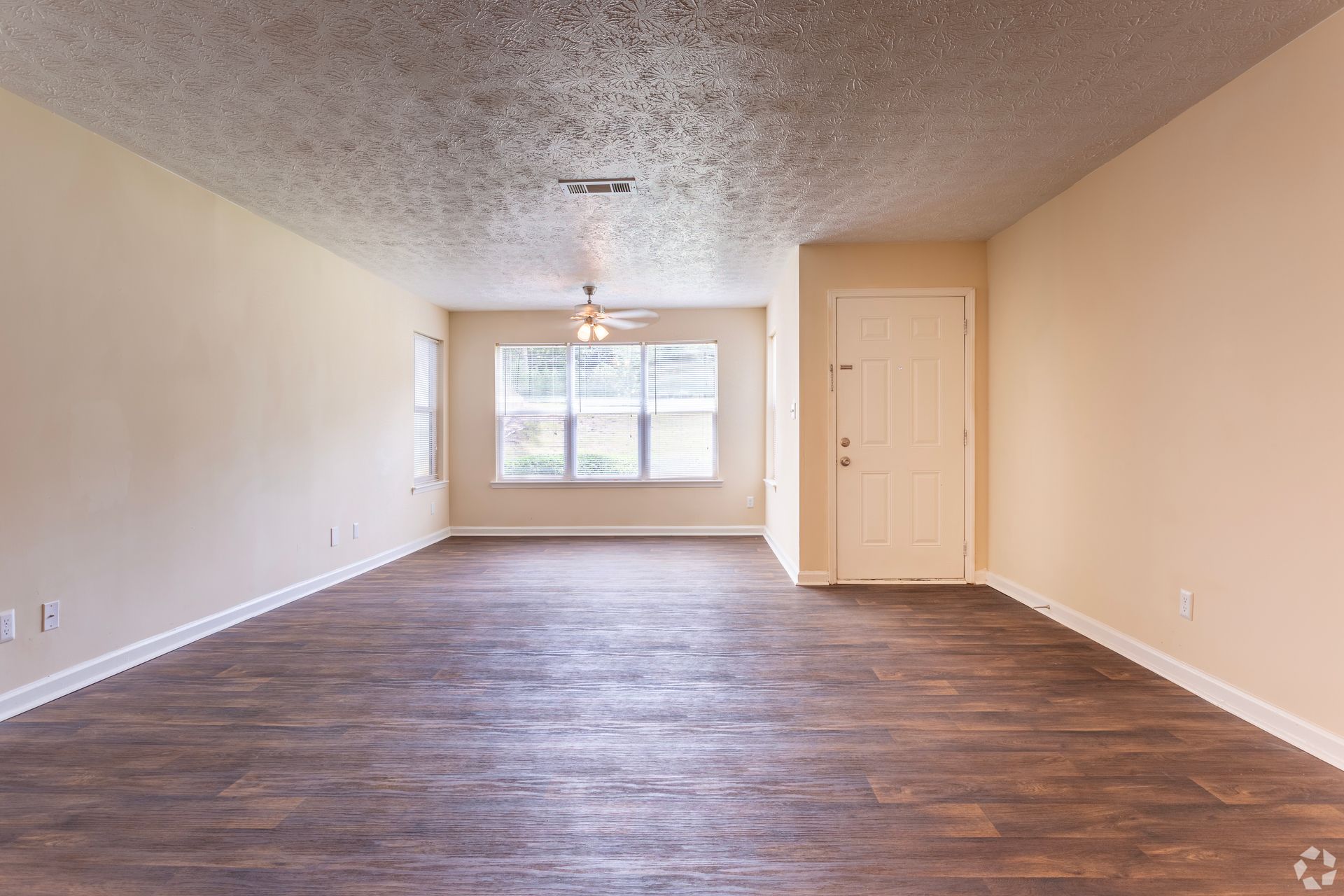 An empty room with light tan walls, a textured ceiling, dark wood flooring, a window, and a white entrance door.