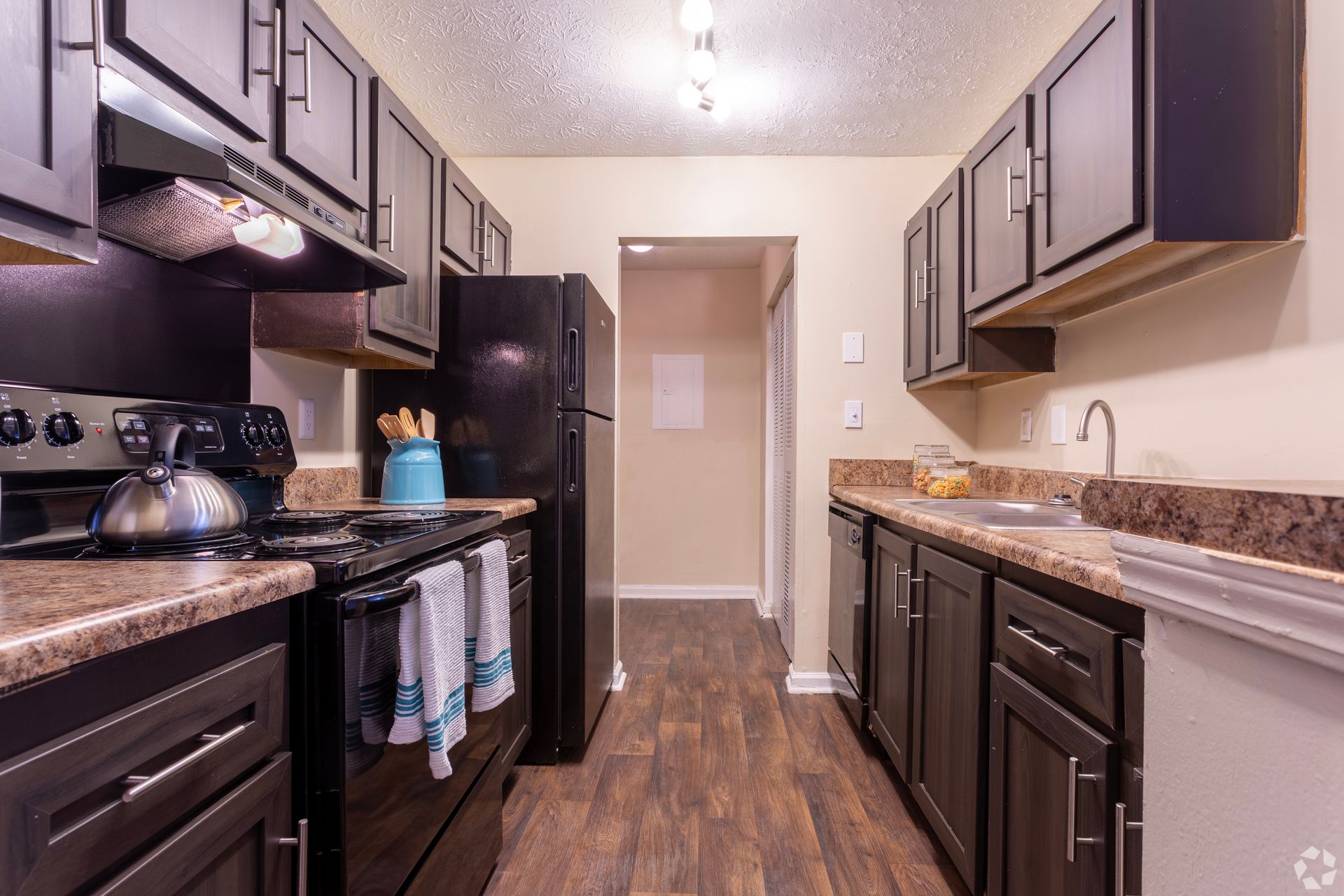 A narrow kitchen with dark cabinets, black appliances, speckled countertops, and wood-look flooring.