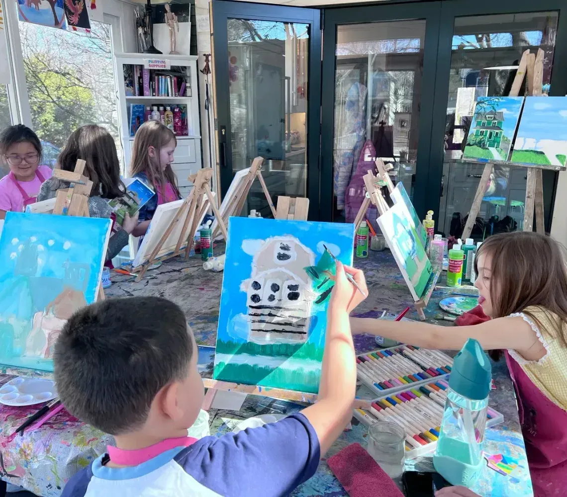 Children sit at tables in a bright, sunlit room, painting landscapes on canvases set on wooden easels.