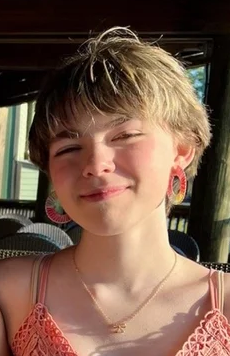 A person with short, light-brown hair smiling, wearing a peach-colored top, a gold necklace, and colorful hoop earrings.