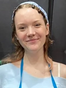 A person with wavy hair, wearing a light blue headband with clips and a blue apron, smiles against a dark background.