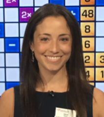 A smiling person stands in front of a game show board with various numbers and symbols in a grid.