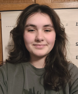 A person with shoulder-length wavy brown hair smiling at the camera, wearing an olive green shirt in an indoor setting.