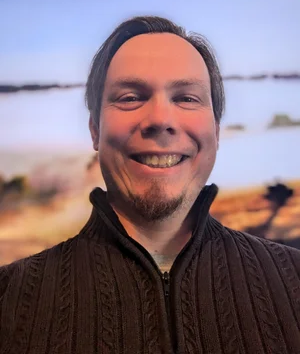 A smiling person with a goatee wearing a dark brown, cable-knit quarter-zip sweater against a blurred outdoor background.
