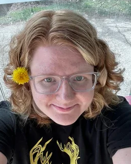 A smiling person with shoulder-length wavy blonde hair wears glasses and a yellow flower tucked behind one ear.