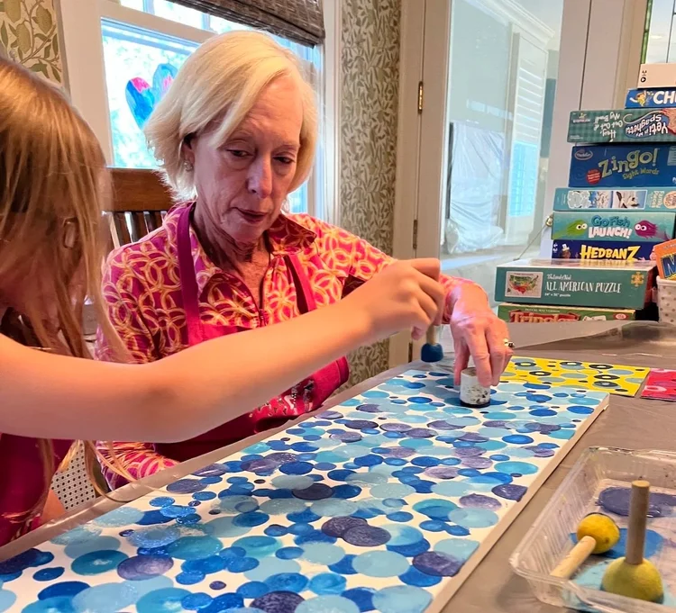 A person and a child work together to paint a long board with blue circles using sponge dabbers at a table.
