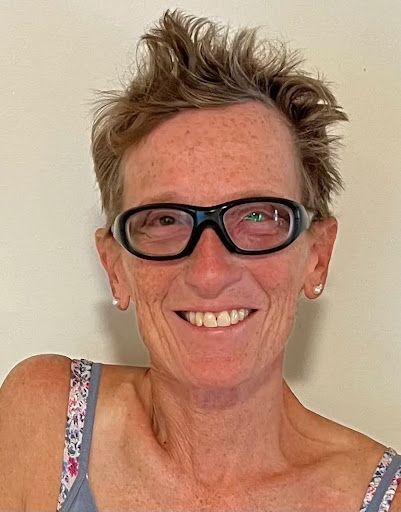 Smiling person wearing black glasses and a floral-strap top against a plain wall