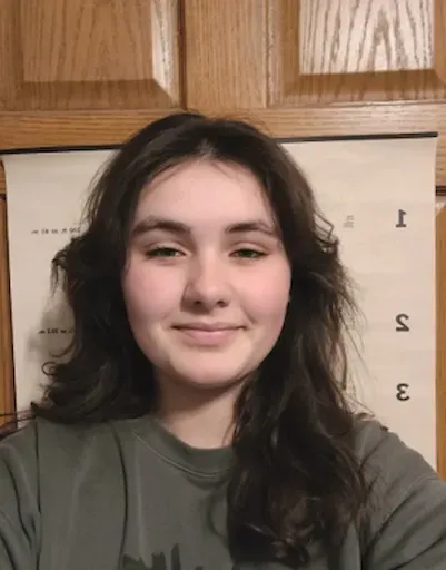 Person with dark wavy hair and a slight smile taking a selfie indoors in front of wooden cabinets.