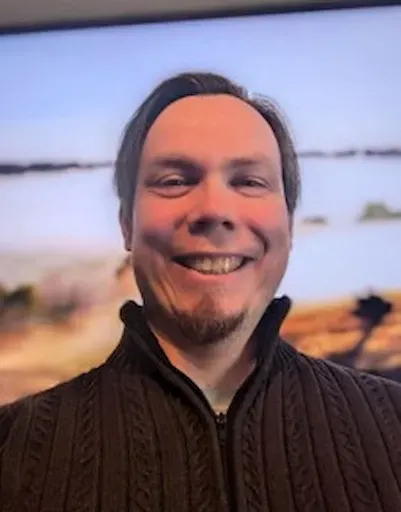 Smiling person in a dark sweater with a blurred lakeside landscape behind them