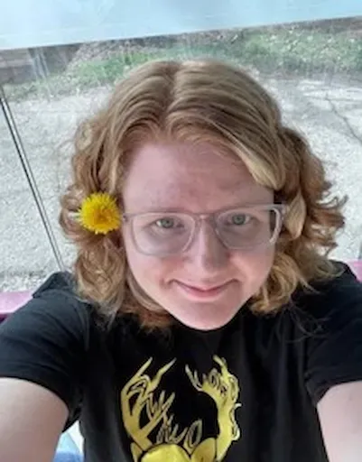 Smiling person with glasses and a yellow flower in their hair, wearing a black shirt, outdoors on a patio.
