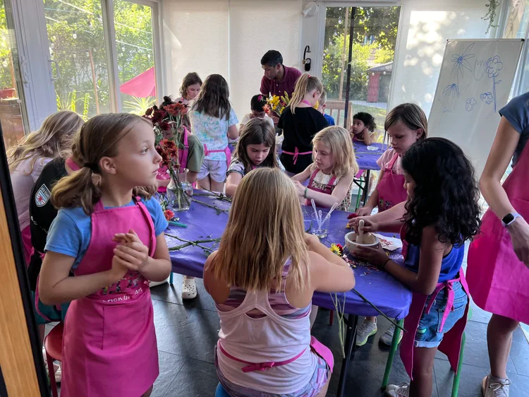 A group of people wearing pink aprons working at a long table with flowers and craft supplies in a bright room.