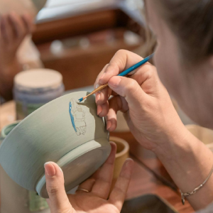 A person paints a small blue line on a light green ceramic bowl with a fine-tipped brush at a table with paint jars.