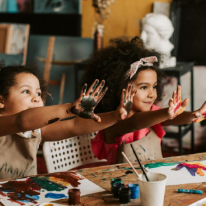 Three children with paint-covered hands pose with outstretched arms in a bright art studio filled with supplies.
