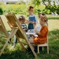 Children painting on easels in a sunny, green park near a building.