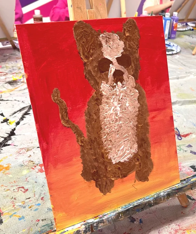 A thick acrylic painting of a brown cat with a white chest on a red and orange background, sitting on an easel.