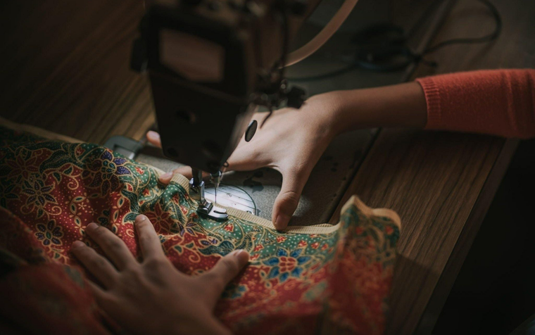 Hands guide a piece of patterned, colorful fabric under the needle of a sewing machine on a wooden surface.