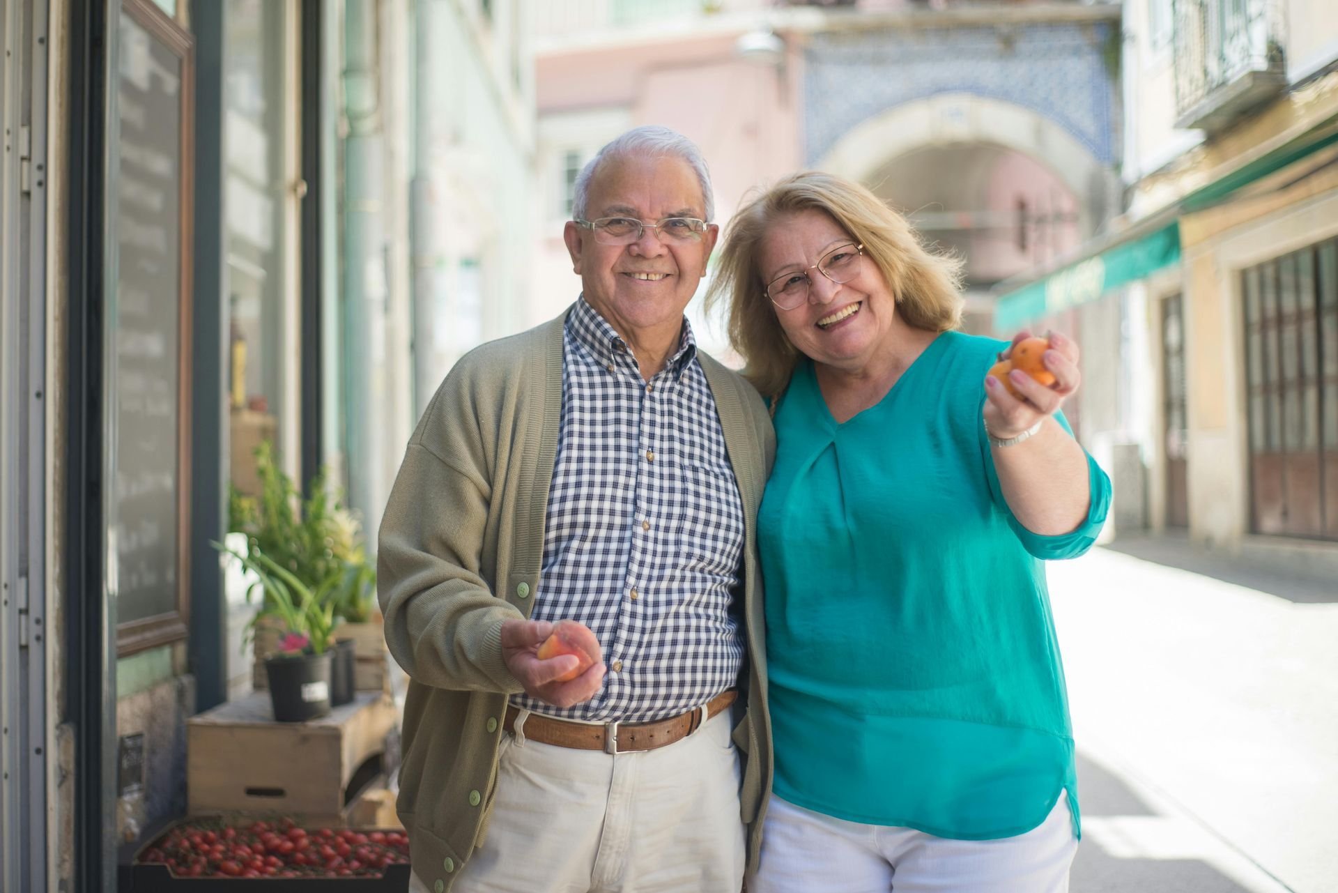 Pareja de ancianos sonriendo, sosteniendo naranjas, de pie en una calle soleada con tiendas.