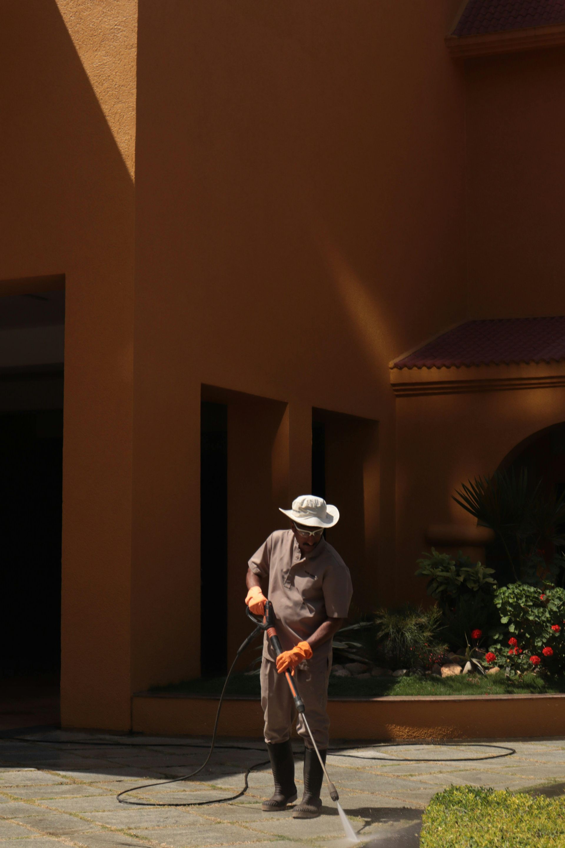 Man wearing a hat and gloves, washes a paved area near an orange building with a water hose.