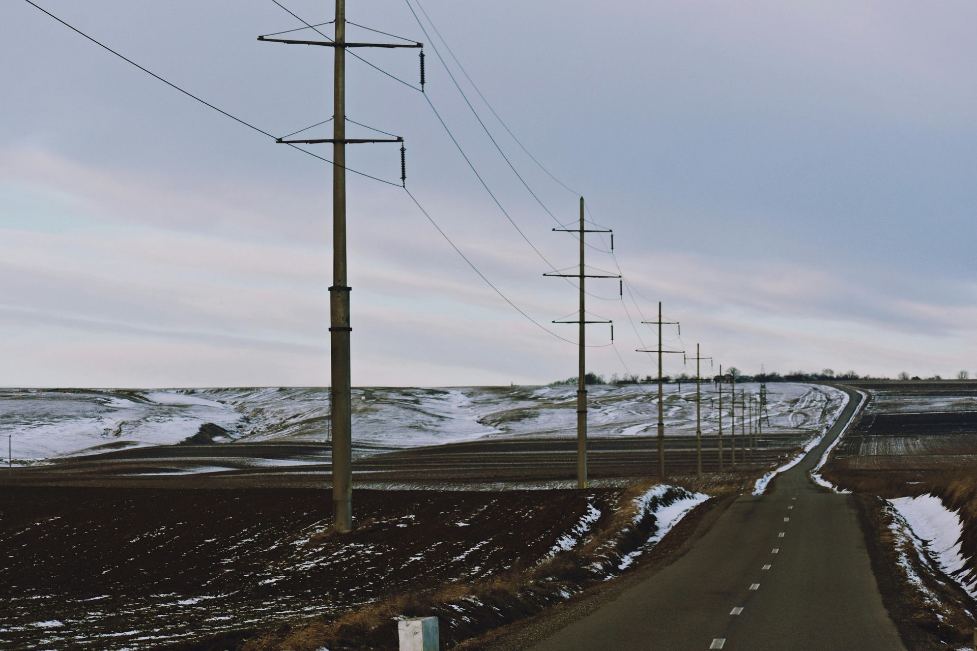 Road through a rural landscape with power lines. Cloudy sky, snow patches on hills and fields.