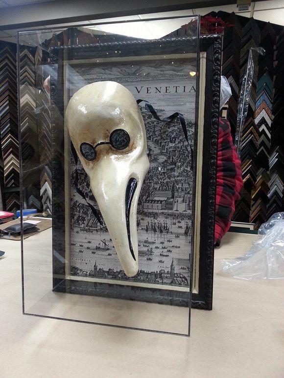 A plague doctor mask displayed in a framed shadow box in front of a black and white map of Venice.