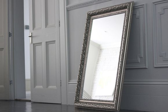 A tall, ornate silver mirror leans against a gray wall near an open white door.