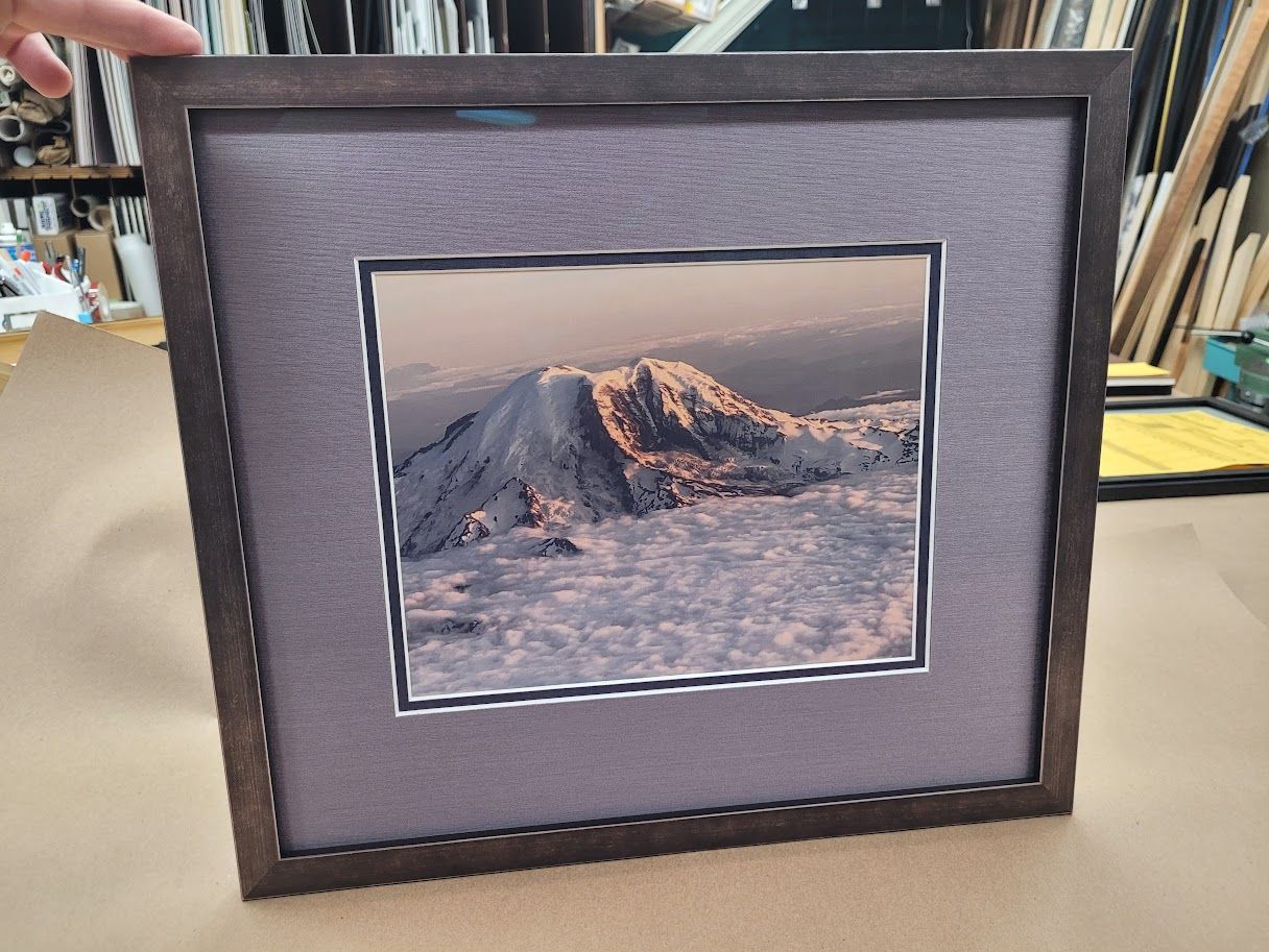 Square framed photo of a snowy mountain peak, surrounded by a gray fabric mat and brown frame.