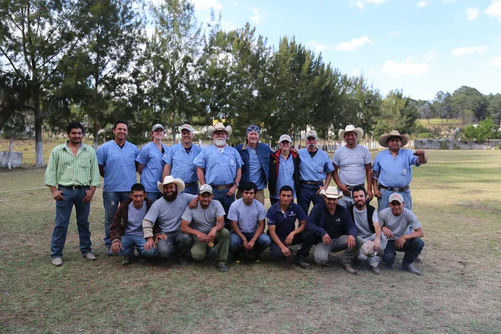 Group of men pose on grass field, some wearing cowboy hats. Trees in background.