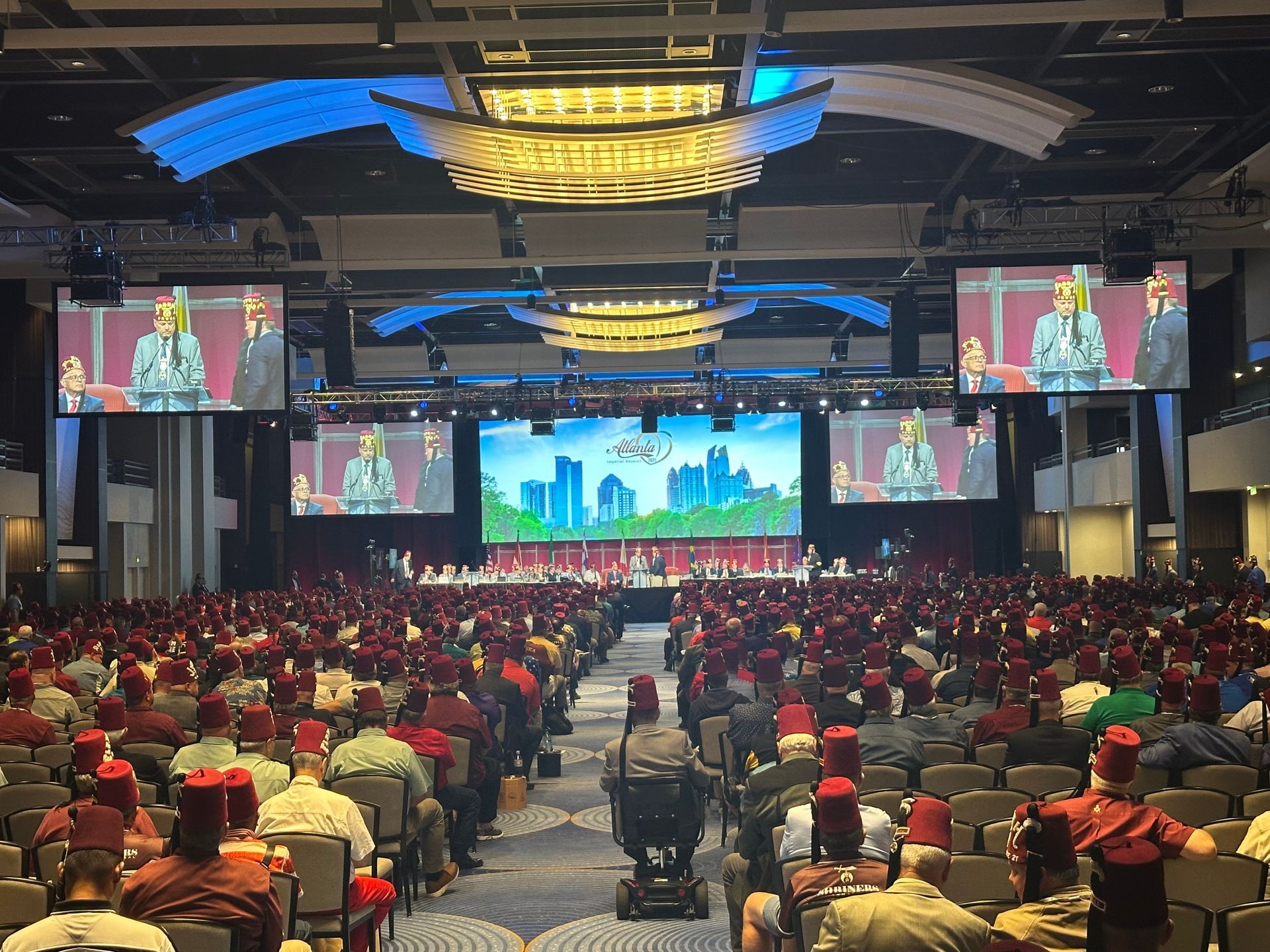 A large crowd wearing red fezzes sits in a convention hall facing a stage with several large screens showing a speaker.