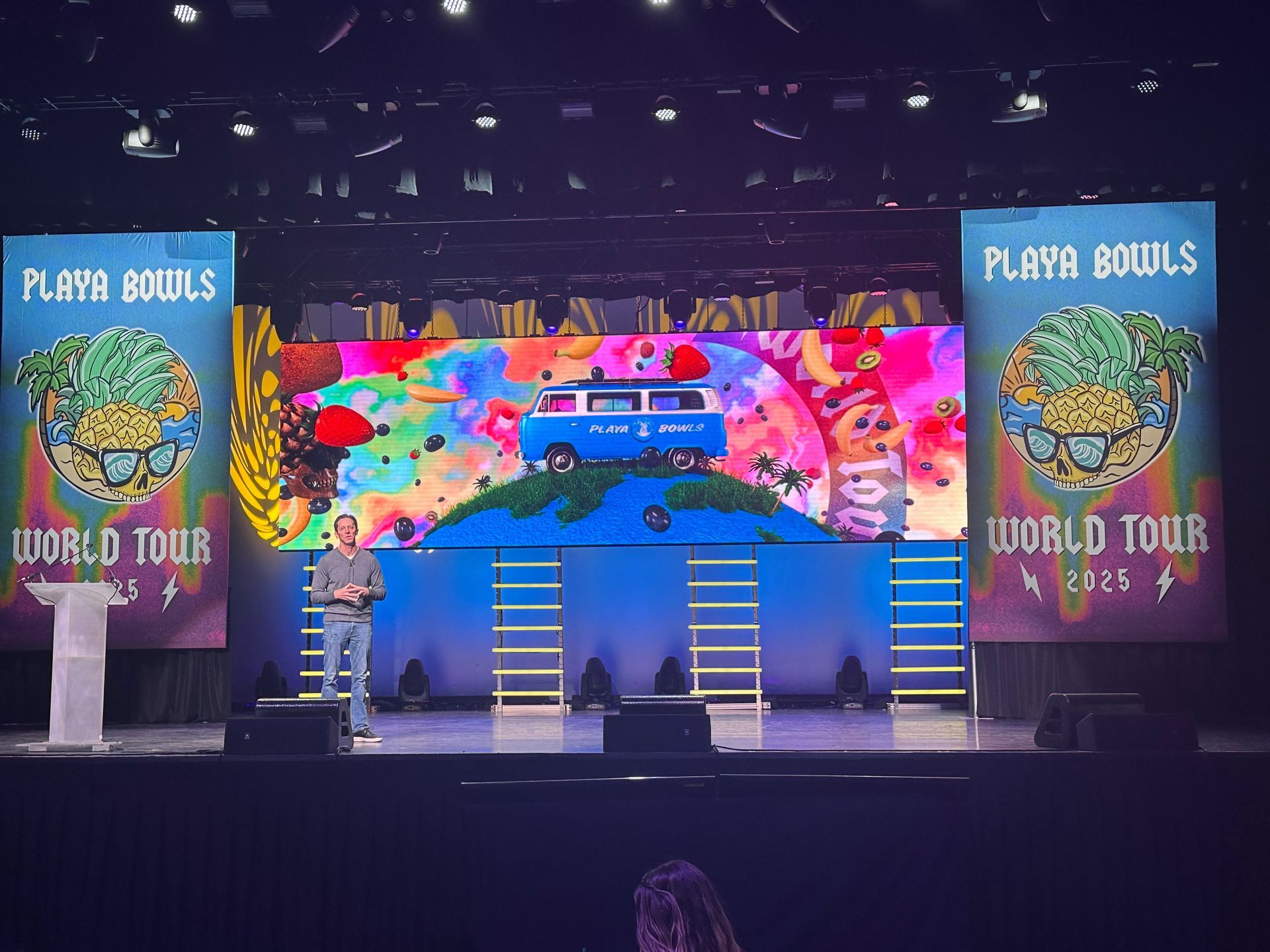 A person standing on a stage at a Playa Bowls World Tour 2025 event featuring colorful banners and a screen with a van.
