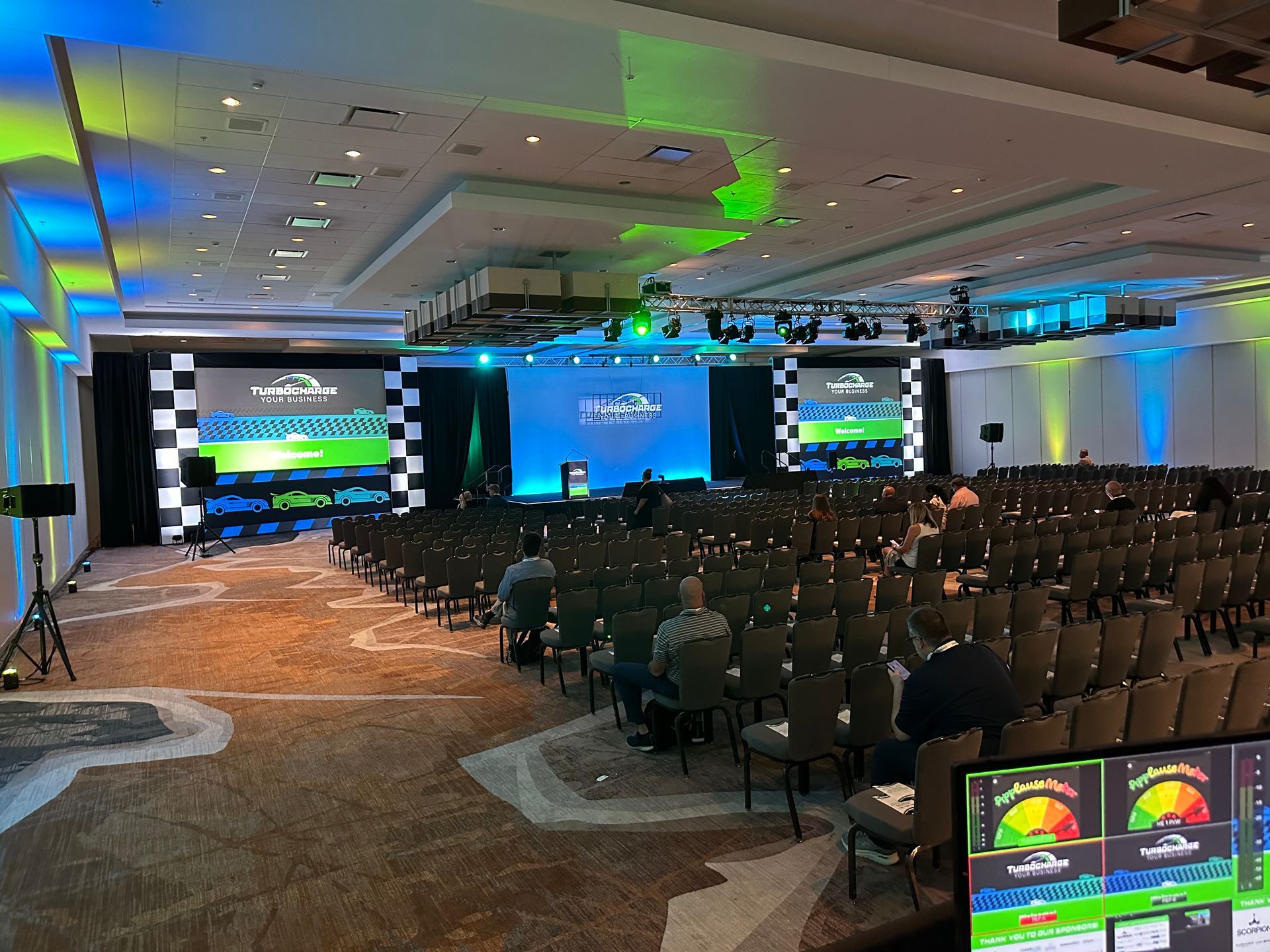 Conference hall with rows of chairs facing a stage featuring large screens, checkered patterns, and blue and green lighting.