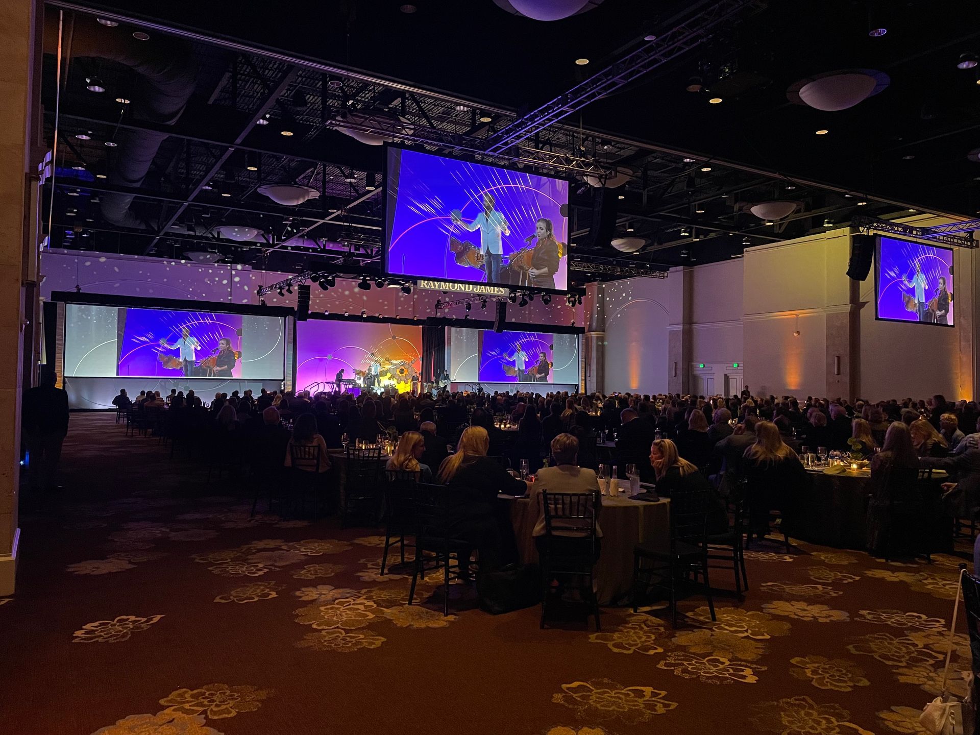 A large audience sits at round tables in a dim hotel ballroom facing screens projecting a speaker on a stage.