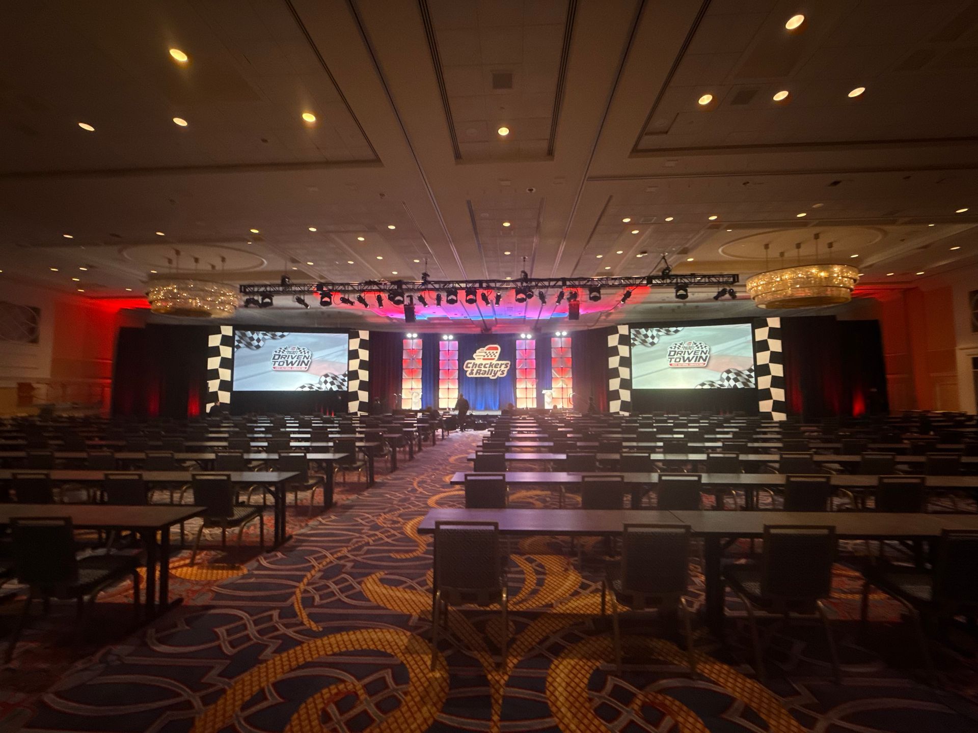 A wide view of a conference hall with rows of tables, a stage featuring two large screens, and vibrant blue-orange lighting.
